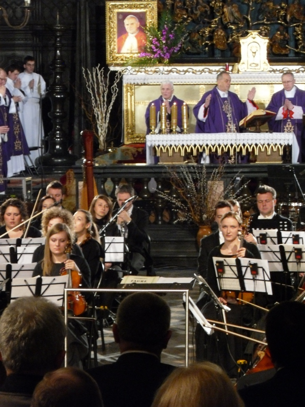 Eine Gruppe von Musikern, die Geigen spielen, mit Notenblättern vor sich, mit drei Kirchenvätern im Hintergrund, einer Bibel vor ihnen und einem gerahmten Bild mit Einlegearbeiten drumherum.