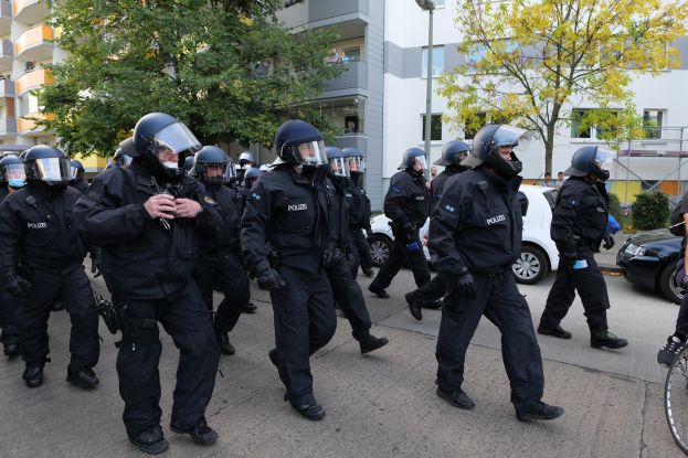 Eine Gruppe von Polizisten in Einsatzausrüstung geht eine Straße entlang, mit einer Person, die ein Fahrrad auf der rechten Seite hält, Fahrzeugen, Bäumen, Gebäuden und einem Laternenpfahl im Hintergrund.