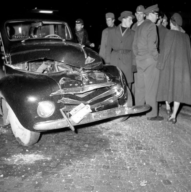 Ein Schwarz-Weiß-Bild eines verbeulten Autos am Straßenrand mit einer Gruppe von Menschen drumherum, die das erhebliche Ausmaß der Beschädigung der Motorhaube und der zerbrochenen Fenster begutachten.