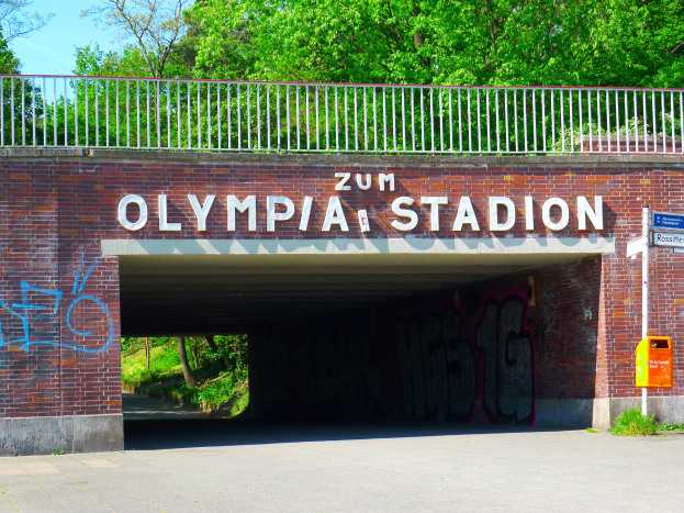 Der Eingang zum Olympiastadion in Berlin, Deutschland, mit einer Brücke mit Text, einem Metallzaun, einem Schild, einer Box, Pflanzen, Gras, Bäumen und einem bewölkten Himmel.