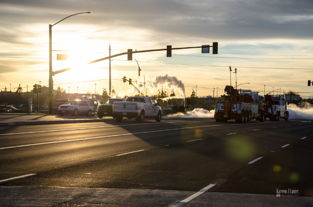 Mehrere Fahrzeuge bewegen sich auf einer Straße mit Verkehrszeichen, Masten und einer Industrieanlage, die Rauch im Hintergrund freisetzt, unter einem sonnigen Himmel.
