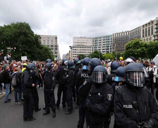 Eine große Gruppe von Polizisten steht vor einer Menge von Menschen auf einer Straße, die von Bäumen und Gebäuden mit Fenstern umgeben ist, unter einem bewölkten Himmel, mit einigen in der Menge, die Kameras halten, während einer Protest in Berlin, Deutschland.