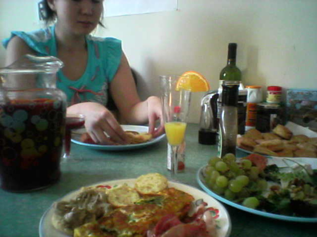 Eine Frau sitzt auf einem Stuhl mit einem Teller voller Essen, darunter Trauben, und verschiedenen Gegenständen wie einem Glas, Flasche und Schachtel in der Nähe.