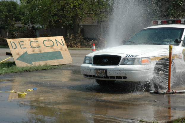 Ein Polizeiwagen fährt durch eine Wasserlache, mit einer Tafel mit Text, einem Verkehrskegel und anderen Gegenständen am Boden; es gibt Bäume, Gebäude und einen klaren blauen Himmel im Hintergrund.