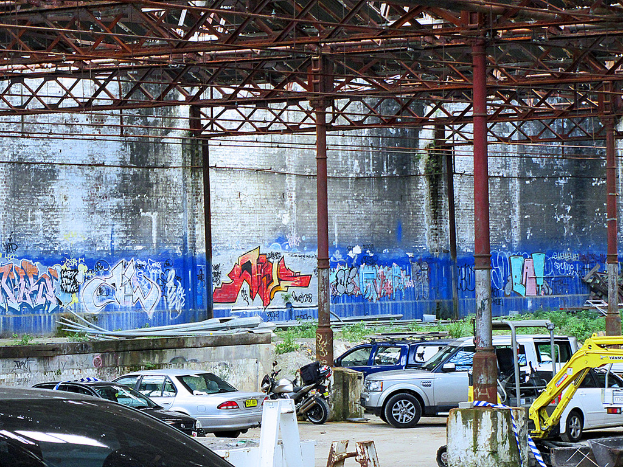 Fahrzeuge, Eisenstangen und Pflanzen am Boden mit einer Warnbinde, Gegenstände, Stangen und eine bemalte Wand im Hintergrund.
