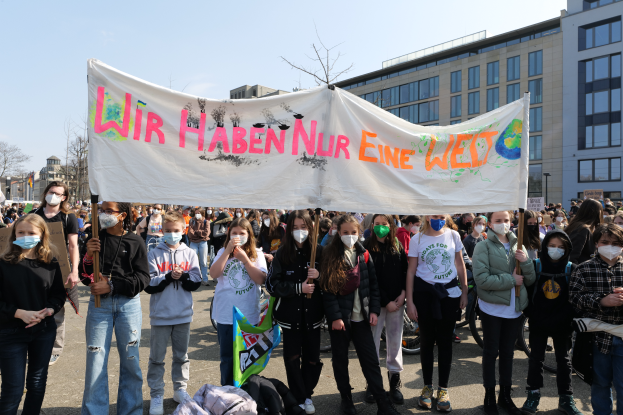 Eine Gruppe von Menschen mit Masken hält ein Transparent mit der Aufschrift "Wir haben nur eine West" vor einem Gebäude mit Fenstern, umgeben von Bäumen und einem klaren Himmel, mit Taschen und anderen Gegenständen, die auf dem Boden verstreut sind, während einer Klimademonstration in Deutschland.