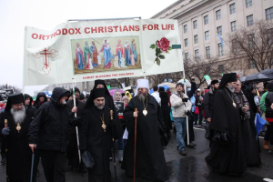 Eine Gruppe orthodoxer Christen marschiert durch eine Straße, hält ein Banner mit Text und Bildern, einige tragen Mützen und halten Stöcke, andere halten Schirme, mit Bäumen, Gebäuden und einem klaren blauen Himmel im Hintergrund.