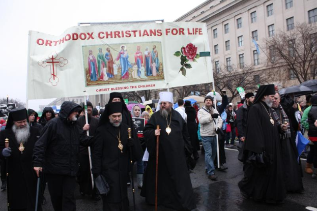 Eine Gruppe orthodoxer Christen marschiert durch eine Straße, hält ein Banner mit Text und Bildern, einige tragen Mützen und halten Stöcke, andere halten Schirme, mit Bäumen, Gebäuden und einem klaren blauen Himmel im Hintergrund.