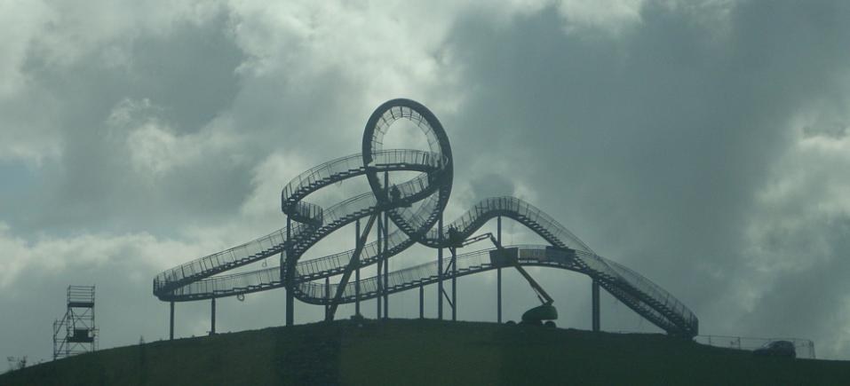 Eine Achterbahn auf dem Höhepunkt eines Hügels mit einem bewölkten Himmel als Hintergrund, umgeben von Stützpfählen und -strukturen.