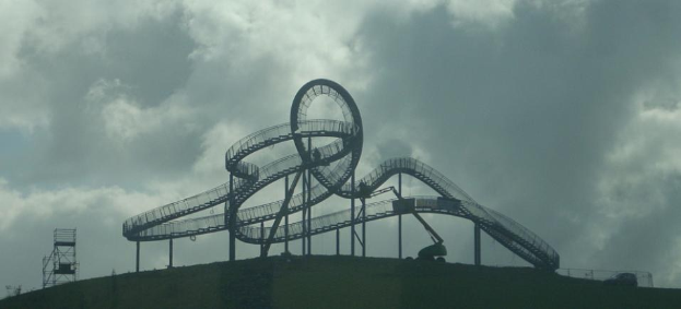 Eine Achterbahn auf dem Höhepunkt eines Hügels mit einem bewölkten Himmel als Hintergrund, umgeben von Stützpfählen und -strukturen.