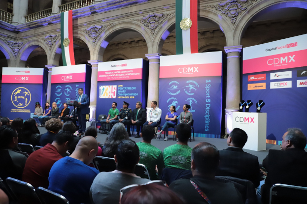 Gruppe von Menschen, die auf Stühlen sitzen und einer Bühne mit einem Mann neben einem Mikrofon auf einem Lautsprecherständer und Event-Bannern mit der Aufschrift "Pentathlon-Weltmeisterschaften 2018" im Hintergrund zugewandt sind.