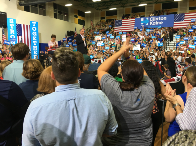 Eine große Menge steht vor einer Bühne, auf der ein Mann in ein Mikrofon spricht, eine Frau steht neben ihm, mit Fahnen, Transparenten und einem Lautsprecher auf der Bühne, und die Menge klatscht und jubelt.