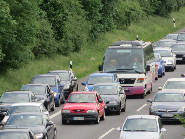 Ein Stau auf einer Autobahn mit vielen Autos, einem Van und Menschen in den Fahrzeugen, mit Bäumen und Gras im Hintergrund.
