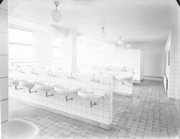 Schwarz-weiß-Foto einer öffentlichen Toilette mit Waschbecken, Wasserhähnen, Wänden, Lampen und Fenstern.