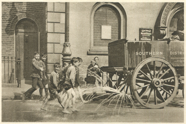 Kinder in Feuerwehruniformen versammeln sich um einen Feuerwehrmann, der einen Helm hält, mit einem Feuerwacheninneren im Hintergrund.