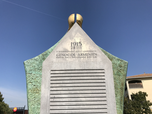 Ein steinernes Denkmal mit einer Uhr darauf, das mit Text beschriftet ist, steht in einem Park umgeben von Bäumen und Gebäuden unter einem klaren Himmel, das den 24. Jahrestag des Völkermords in Armenien gedenkt.