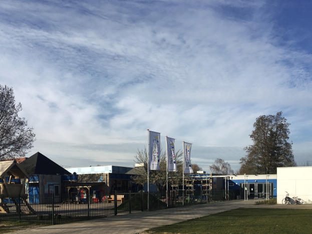 Ein großes Gebäude mit einem grasbewachsenen Bereich davor, umgeben von einem Metallzaun, einem Weg, Bäumen, Pfosten, Bannern, einem geparkten Fahrrad, nahen Häusern und einem bewölkten Himmel, das als neuer Spielbereich für Kinder an einer Schule dient.