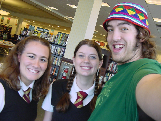 Ein Mann und zwei Schüler machen ein Selfie in einer Bibliothek, im Hintergrund sind Bücherregale mit vielen Büchern zu sehen.