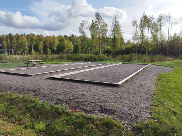 Eine grüne Wiese mit einer Picknick-Tafel, umgeben von Bäumen unter einem klaren blauen Himmel, mit Spielplatz-Geräten wie Stangen und einer Bank links.