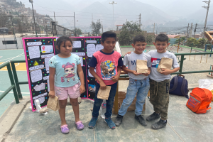 Eine Gruppe von Kindern steht auf einem Basketballplatz, jedes hält einen Papiertüte, mit Tüten und Gegenständen rechts und einer Texttafel links, mit Zaun, Pfosten, Straßenlaternen, Bäumen, Gebäuden, Hügeln und Himmel im Hintergrund.