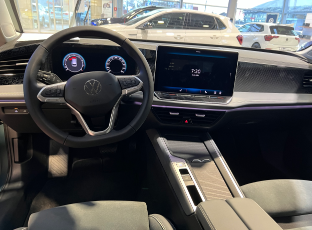 Das Innere eines Volkswagen e-Golf, mit einem Lenkrad, Armaturenbrett, Bildschirmen, Knöpfen, Sitzen und Glastüren, mit einer Aussicht auf andere Autos, Gebäude, Bäume, Pfosten und den Himmel durch die Glastüren.