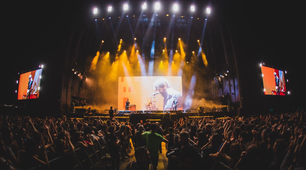 Eine große Menschenmenge vor einer hell erleuchteten Bühne mit Musikern und einem Sänger mit Mikrofon, vor einem dunklen Hintergrund mit Bildschirmen.