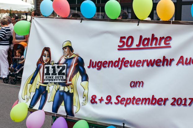 Eine Gruppe von Menschen auf der Straße, einige mit Kinderwagen, mit einem Banner mit dem Text "50 Jahre Jugendfeuerwehr August 2017" und Cartoon-Bildern, einem Zaun, Luftballons, einem Zelt, einem Gebäude mit Fenstern und Bäumen im Hintergrund.