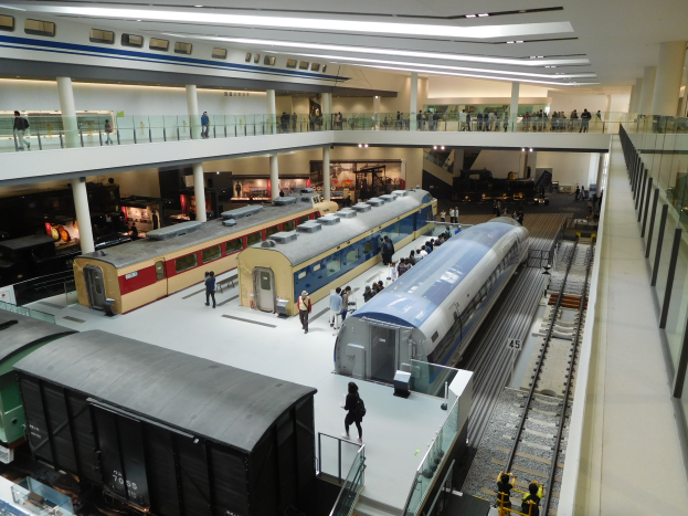 Ein geräumiger Museumsraum voller verschiedener Züge zur Besichtigung, Menschen, Geländer, Säulen und Deckenleuchten.