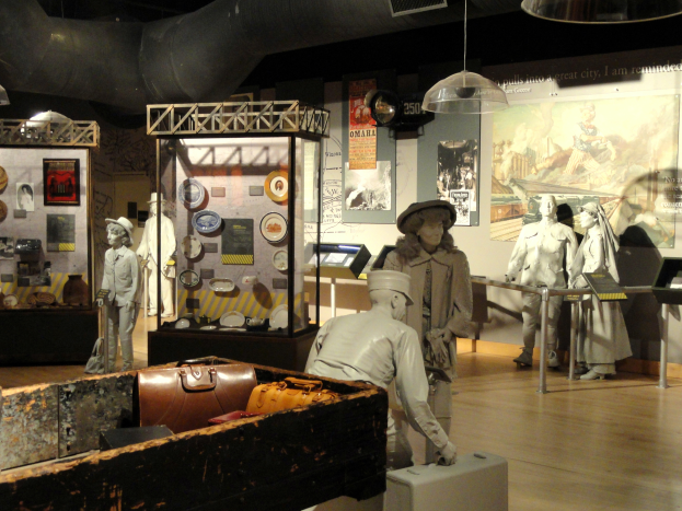 Ein Museumsraum mit verschiedenen Ausstellungsstücken, darunter Puppen mit Hüten, eine Aktentasche, eine Kiste und andere Gegenstände auf dem Boden sowie Informationsschilder an den Wänden und Lampen an der Decke.