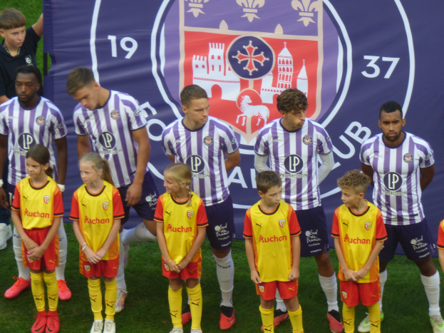 Eine Gruppe junger Menschen, die auf einem Fußballfeld zusammenstehen und ein Banner mit der Aufschrift "Preston Primary School Football Team" im Hintergrund halten.