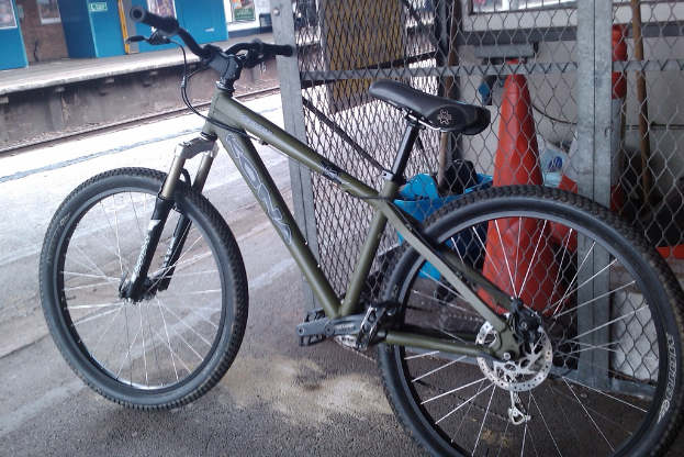 Ein grünes Fahrrad steht im Vordergrund vor einem Zaun, einer Verkehrskegel, einer Straße, einem Bahnsteig und einer Bahnstrecke.