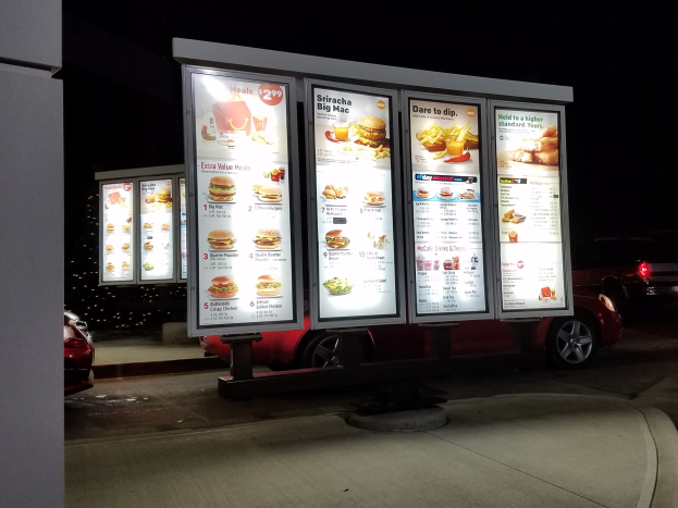 Fast food restaurant bei Nacht mit beleuchteter Reklame, fahrende Fahrzeuge im Hintergrund, eine Säule links und ein dunkler Himmel.