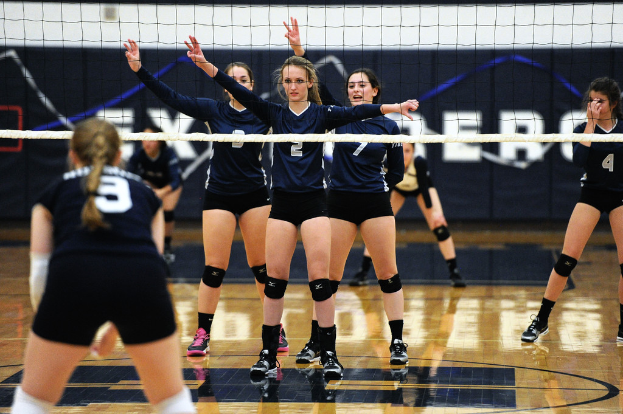Drei Mädchen bei einem Volleyballspiel auf dem Feld mit erhobenen Händen, ein Netz vor ihnen und ein Zaun im Hintergrund.