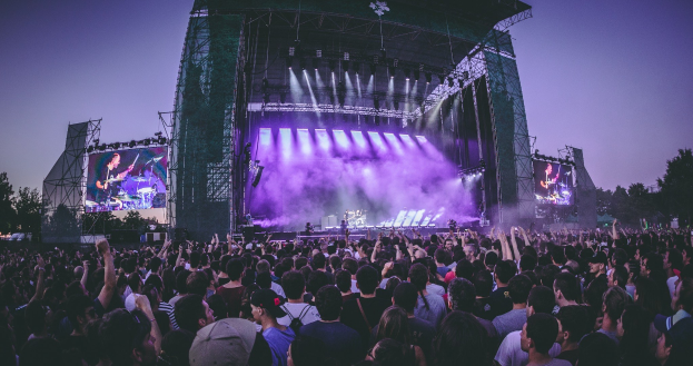 Eine große Menschenmenge bei einem Musikfestival schaut Künstlern auf einer hell erleuchteten Bühne mit Bildschirmen zu, die vor einem Gebäude, Bäumen und einem klaren blauen Himmel aufgebaut ist.