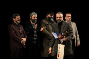 Eine Gruppe von Männern steht auf einer Bühne, einer hölt eine Tasche und eine Trophäe, mit einem dunklen Hintergrund, der die Eröffnung der Berliner Filmfestspiele suggeriert.