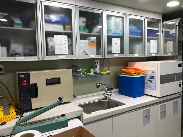 Ein Labor mit einem Waschbecken, einem Wasserhahn, einer Mikrowelle, Drähten, einem Karton und anderen Gegenständen auf der Arbeitsplatte, Schränken mit Papieren daran und Lampen an der Decke.