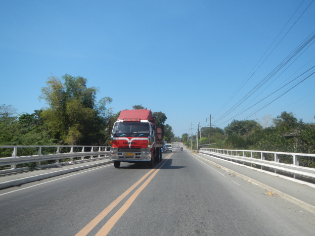 Ein roter Lastwagen fährt auf einer Autobahn mit Leitplanken, Bäumen und Strommasten unter einem klaren blauen Himmel.