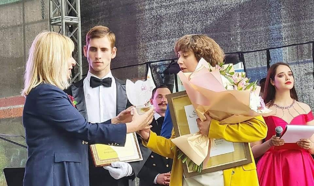 Gruppe von Menschen auf einer Bühne, einer in gelbem Kleid mit Blumen, ein anderer mit Papieren und Mikrofon, mit einer Person links im blauen Blazer und weißem Hemd, die einen Preis entgegennimmt.