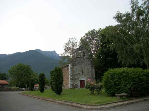 Alte Kirche in einer Straßensituation, umgeben von Häusern, Grün, einer Bank und einer Straße, mit Hügeln und einem blauen Himmel im Hintergrund.