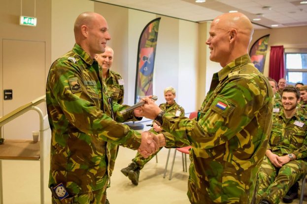 Zwei Männer in militärischer Uniform schütteln sich die Hände vor einer sitzenden Gruppe, mit einem Tisch, Geländer, Fahnen, einer Tür, einem Schild, einem Fenster mit Vorhang und Deckenleuchten im Hintergrund.