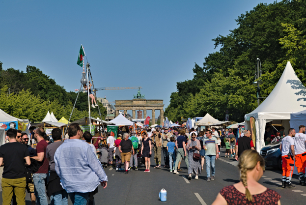 Eine Menschenmenge, die eine Straße mit Zelten, Fahrzeugen und Bäumen entlanggeht, mit einem Bogen und einem klaren blauen Himmel im Hintergrund und Polen mit Flaggen auf der linken Seite, wahrscheinlich das Oktoberfest in München, Deutschland.