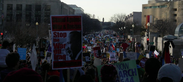 Menschen demonstrieren auf einer Straße und halten Schilder in die Höhe, mit Gebäuden, Bäumen, Laternen und dem Himmel im Hintergrund.