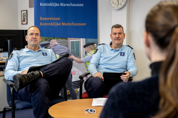 Zwei Polizeibeamte sitzen an einem Tisch mit einer Frau, umgeben von Papieren und Gegenständen, mit einer Fahne, einer Uhr und Foto Rahmen an der Wand und einem weiteren Tisch mit einem Monitor und Zubehör links.