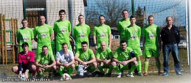 Eine Gruppe von Männern in grünen T-Shirts steht auf einem Fußballfeld, einige in der Hocke, mit einem Ball und einem Torpfosten, umgeben von Bäumen, Gebäuden und Fahrzeugen.