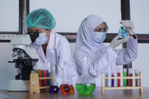 Zwei Frauen in Laborkitteln, Masken, Handschuhen und Hauben arbeiten in einem Labor, wobei eine einen Kolben, Reagenzgläser und andere Gegenstände auf dem Tisch hält und eine Schalttafel im Hintergrund zu sehen ist.