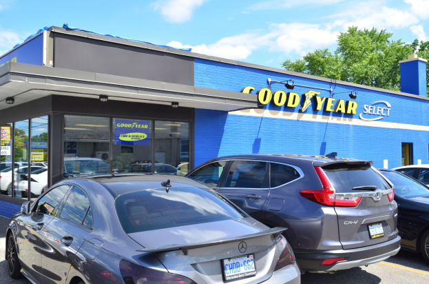 Ein Goodyear Select-Autohaus mit parkenden Fahrzeugen vorne, Beschriftung am Gebäude, Bäumen im Hintergrund und einem bewölkten Himmel.