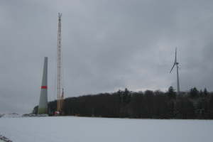 Eine Windkraftanlage steht in einem verschneiten Feld umgeben von Bäumen und Türmen unter einem bewölkten Himmel.