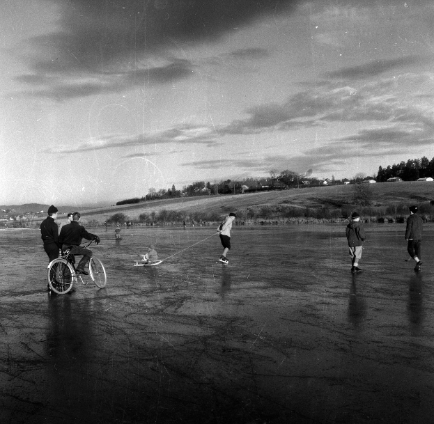 Eine Gruppe von Menschen, die auf einem zugefrorenen See Schlittschuh fährt, mit einer Person, die im Vordergrund Fahrrad fährt, Bäume, Häuser und einen klaren Himmel im Hintergrund, in Schwarz-Weiß dargestellt.