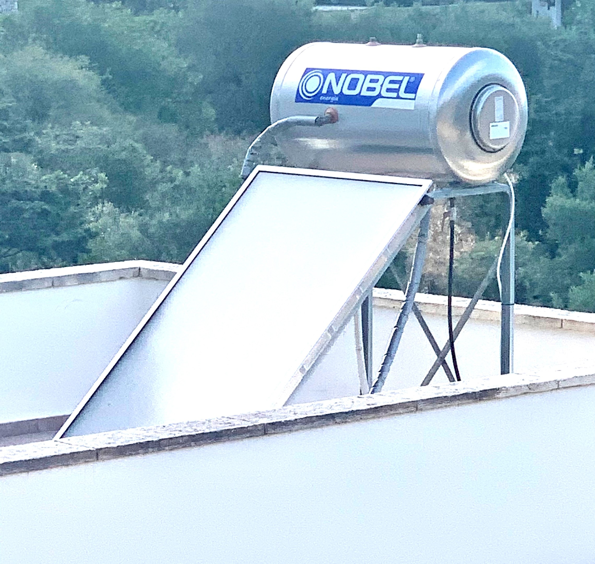 Solarwassererwärmer auf dem Dach eines Gebäudes mit einem angebauten Speicher, vor einem Hintergrund aus Bäumen.