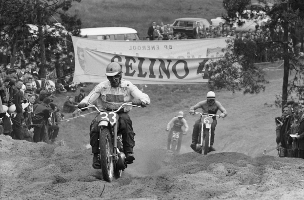 Schwarzes und weißes Bild von Menschen, die auf Dirt Bikes auf einem Dirt Field fahren, mit Zuschauern, einem Banner, Bäumen und Fahrzeugen im Hintergrund.